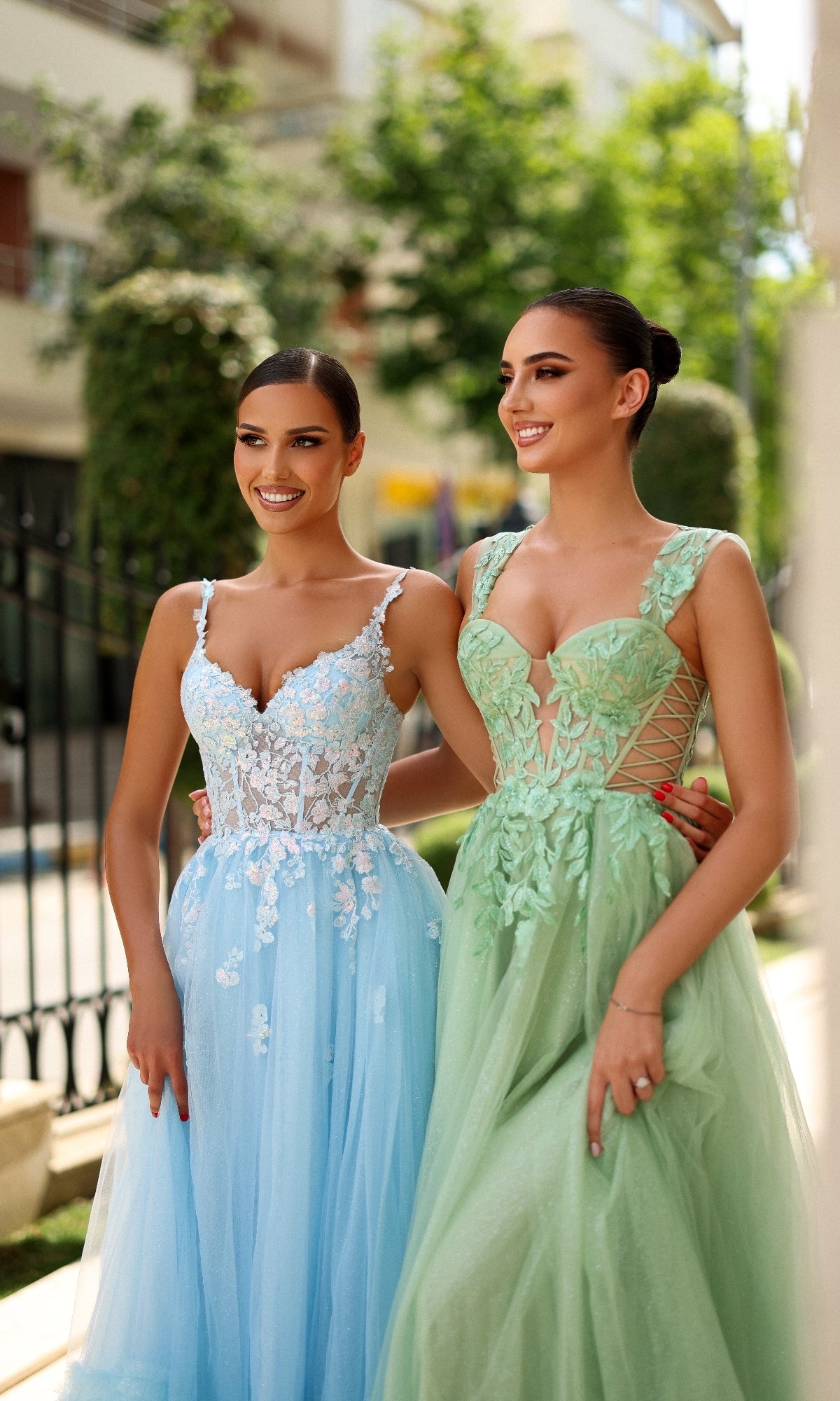 Two women stand outdoors, smiling in elegant sleeveless gowns. One wears the Tina Holly TY405 long formal dress in pastel blue with a tulle A-line skirt and floral details; the other is in light green with a lace corset bodice.